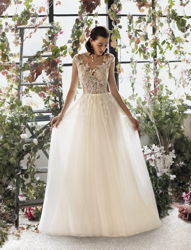 Schwarzhaarige Braut in Brautkleid Vintage lange durchsichtige Ärmel mit geschlossenen Augen in Raum mit weißen und rosanen Blumen sowie Blättern und Kleid zwischen den Fingern