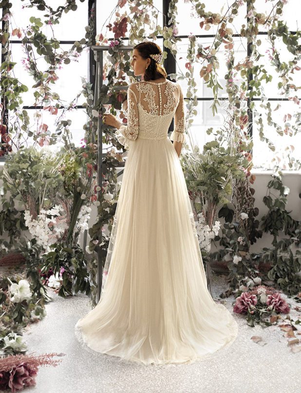 Schwarzhaarige Braut in Brautkleid A-Linie lange Ärmel in Raum mit weißen und rosanen Blumen sowie Blättern Rückenansicht und Kleid zwischen den Fingern