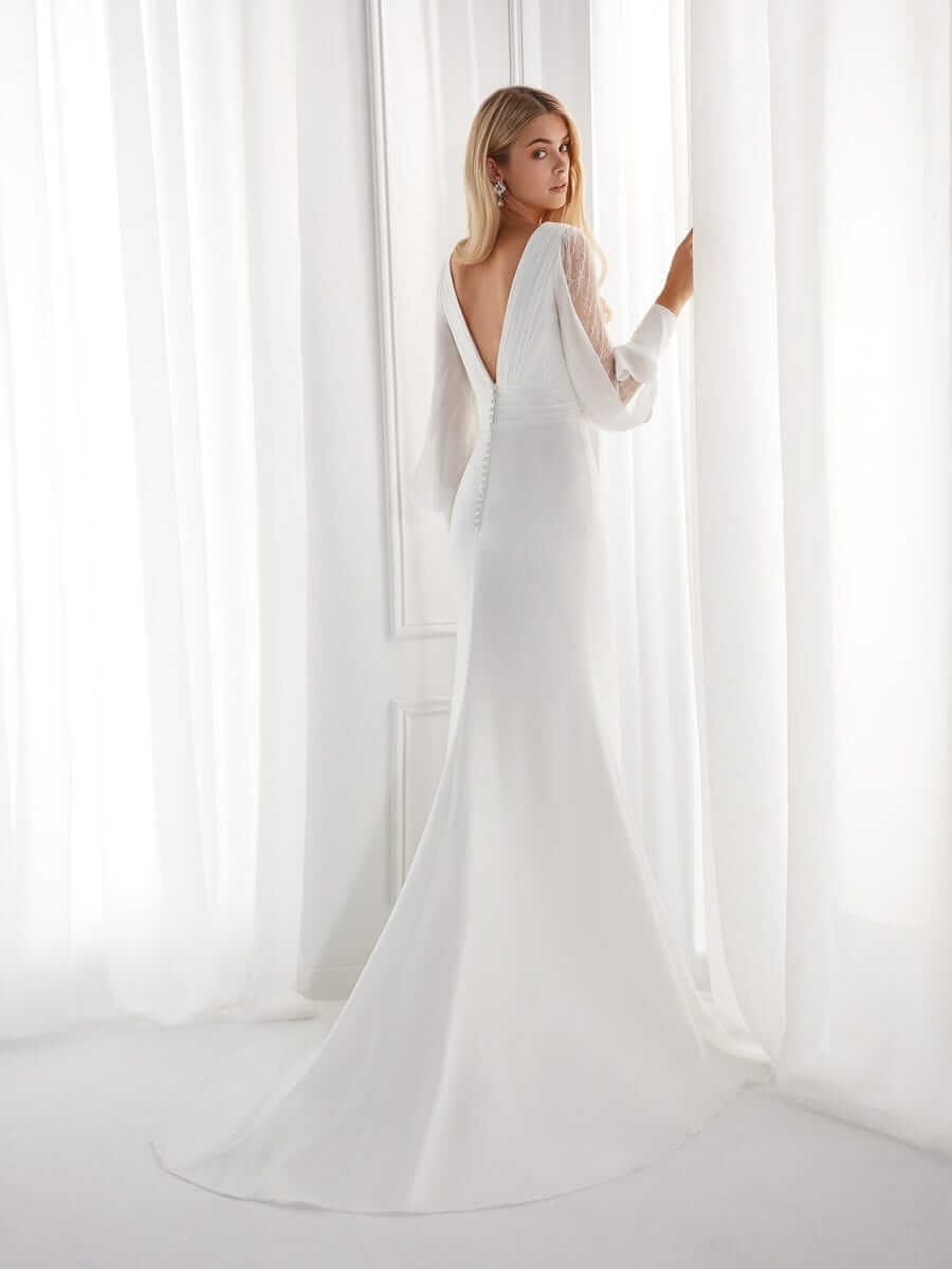 Blonde Braut in Brautkleid Fit & Flare an der Gardine in einem weißen Zimmer mit V-Ausschnitt am Rücken und Schulterblick