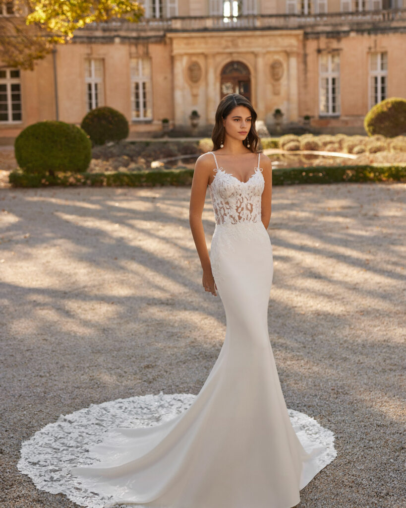 Brünette Frau mit langen offenen Haaren und langen Perlenohrringen trägt ein romantisches und figurbetontes Hochzeitskleid. Das Brautkleid ist ein Kleid, das mit seinem tief ausgeschnittenen Oberteil aus floraler Spitze und seiner eleganten, figurbetonten Silhouette aus Krepp mühelos Charme versprüht. Zarte Träger verleihen diesem auffälligen, figurschmeichelnden Design einen Hauch von Feminität so wie die Kappellenschleppe die einen abschließenden Rand aus Spitze hat. Sie steht im Vorgarten eines Schlosses und steht auf einem Kiesboden.