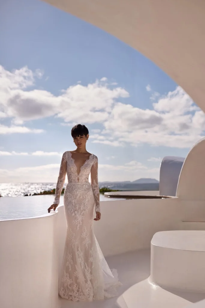 Raffiniertes langärmeliges Brautkleid in Meerjungfrauen-Silhouette aus glitzernder Chantilly-Spitze. Mit tiefem V-Ausschnitt, verziert mit Ornamenten und Pailletten, halbtransparenten, verzierten langen Ärmeln und einem säulenförmigen Spitzenrock mit traumhafter Tüllüberlagerung und einer Midi-Schleppe aus Tüll. Der Rücken besticht durch einen extrem tiefen V-Ausschnitt, der von kunstvoller Spitze umrahmt und mit zarten Knöpfen geschlossen wird und einen atemberaubenden und romantischen Blickfang bildet.