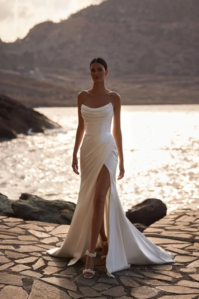 Glamouröses und elegantes A-Linien-Brautkleid aus Satin mit eleganter Drapierung. Das ärmellose Design verfügt über ein strukturiertes Korsett mit sichtbaren Stäbchen, einen kunstvoll drapierten Ausschnitt und einen eng anliegenden Maxirock mit gewagtem Beinschlitz. Seitliche Drapierungen betonen die Silhouette, führen zu einem gewagten Seitenschlitz und enden in einer dezenten Schleppe. Ergänzt werden kann das Kleid durch einen schicken, schulterfreien Bolero Bellissima aus Satin mit langen Ärmeln. Sie können auch eine abnehmbare Schleppe wählen, um das Kleid zu verlängern. Das Kleid verfügt über abnehmbare, schulterfreie Ärmel.