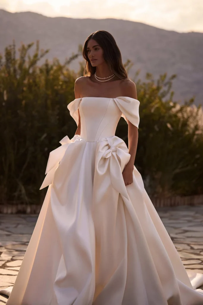 Raffiniertes A-Linien-Brautkleid aus glänzendem Satin. Mit Korsett-Oberteil mit taillierter Taille, drapiertem Herzausschnitt, Reißverschluss am Rücken und üppigem, voluminösem Rock. Während das Korsett die Silhouette formt, verleiht der Rock dem Design mit seinem leichten Volumen einen majestätischen, fließenden Touch. Der Look kann mit Orlena-Handschuhen abgerundet werden. Abnehmbare Off-Shoulder-Ärmel und Träger sind im Preis inbegriffen.