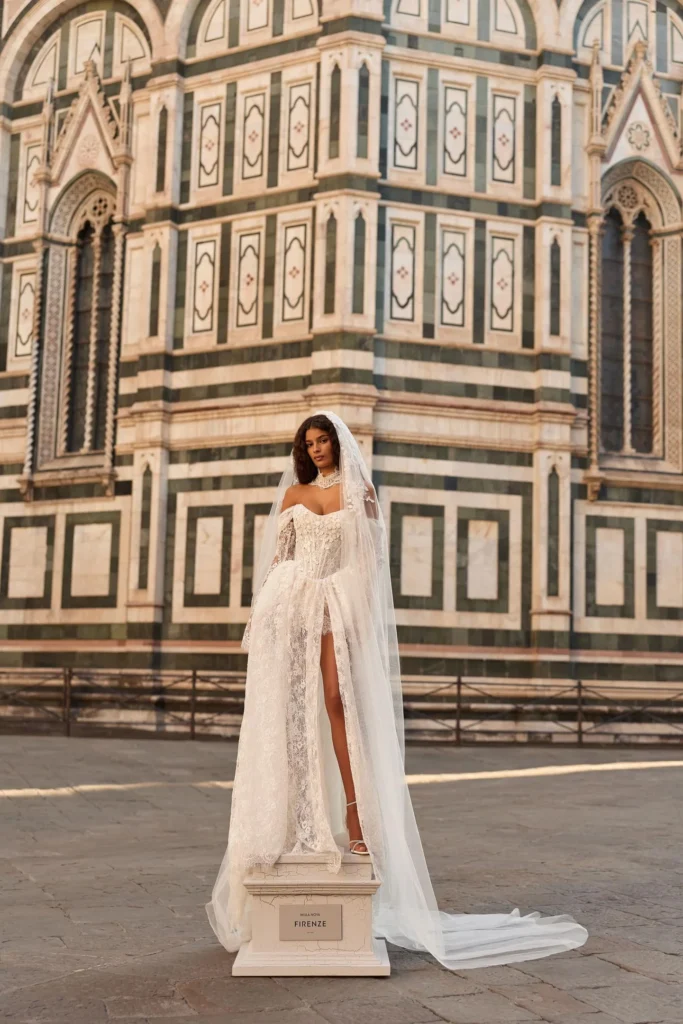Braut in elegantem Spitzen-Brautkleid mit Schlitz und langem Schleier, stehend vor kunstvoller italienischer Kathedralfassade