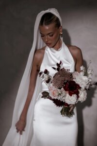 Braut im eleganten Satin-Brautkleid mit Neckholder-Ausschnitt und Schleier, hält einen Brautstrauß in Bordeaux- und Creme-Tönen