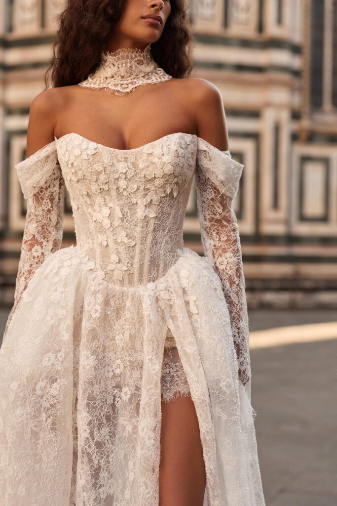 Detailaufnahme eines modernen Brautkleids aus Spitze mit Off-Shoulder-Trägern, transparentem Korsagen-Oberteil, langen Ärmeln und hohem Beinschlitz.