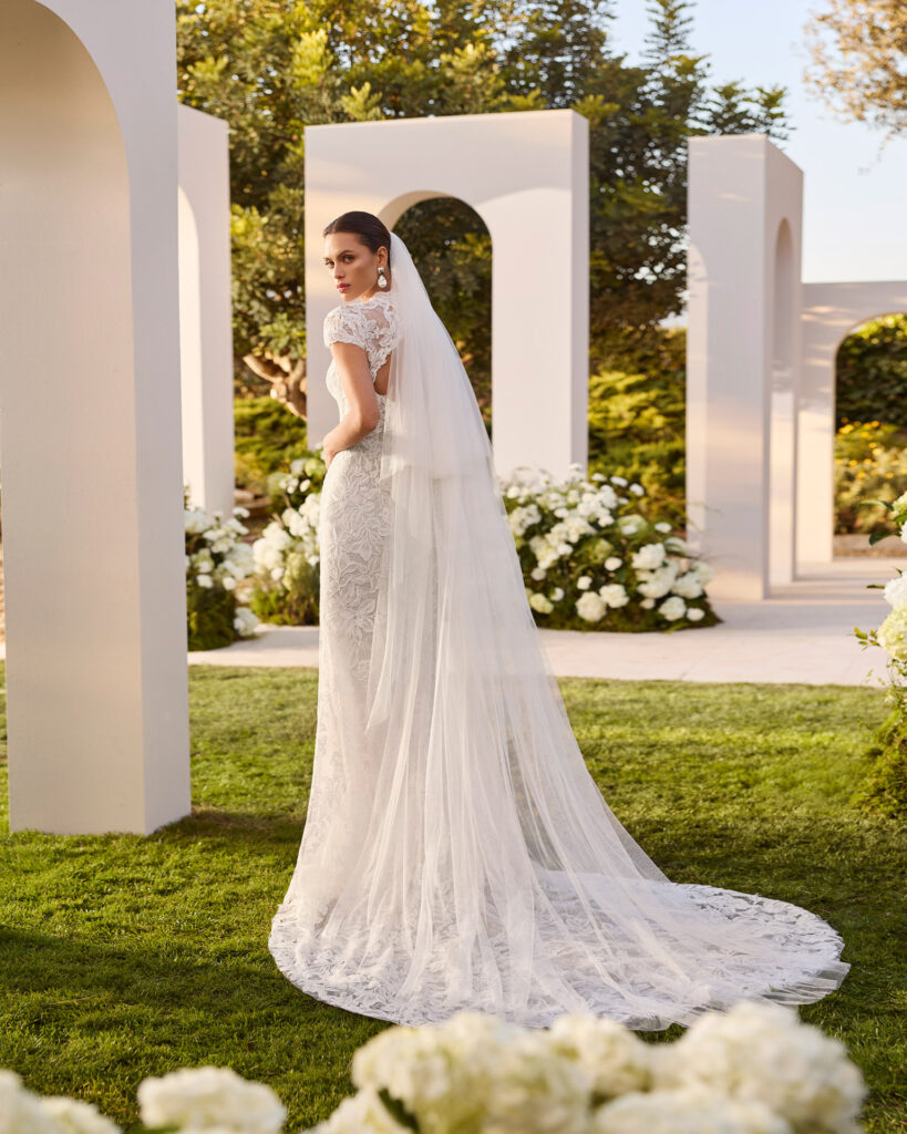 Rückansicht eines romantischen Spitzenbrautkleides mit transparentem Schleier, floraler Spitze und fließender Schleppe vor einer stilvollen Outdoor-Kulisse mit weißen Rundbögen.
