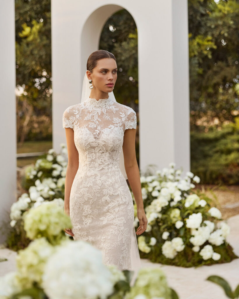 Detailaufnahme eines Brautkleides mit hochgeschlossenem Spitzenoberteil, floralen Applikationen und körpernaher Silhouette, umgeben von weißen Blütenarrangements.