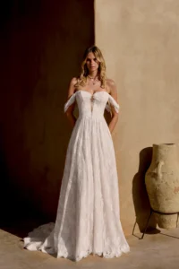 A-Linien-Schnitt aus Blumenspitze mit Off-Shoulder-Ärmeln und tiefem V-Ausschnitt, Frontalansicht vor warmem Lehmwand-Hintergrund.