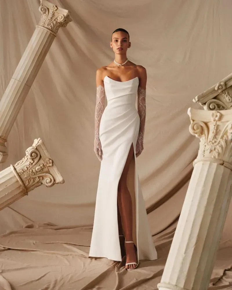 Figurbetontes Crepe-Brautkleid mit Herzausschnitt, Beinschlitz und langen Spitzenhandschuhen, Ganzkörperansicht vor antikem Säulen-Ambiente.