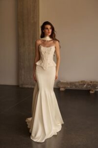 Zweiteiliges Braut-Outfit aus transparentem Spitzenkorsett mit floralen Applikationen und Peplum, kombiniert mit Satin-Meerjungfrauenrock und Choker, Frontalansicht.