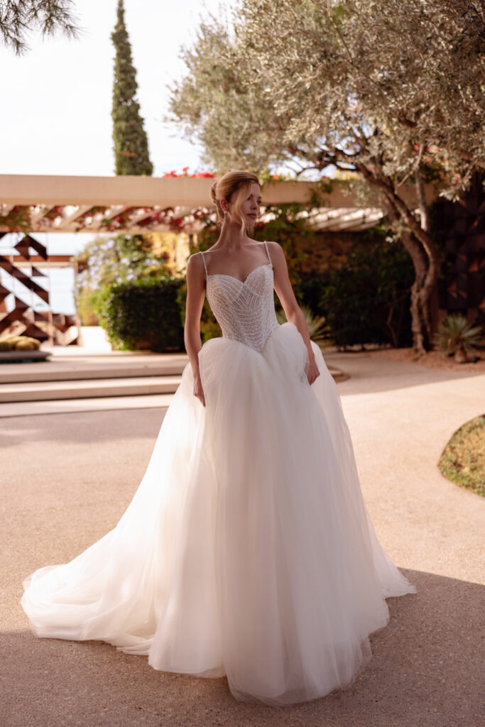 Prinzessinnen-Brautkleid mit Herzausschnitt, dünnen Perlenträgern und besticktem Korsett, kombiniert mit voluminösem Tüllrock und langer Schleppe, Frontalansicht vor mediterranem Gartenambiente.