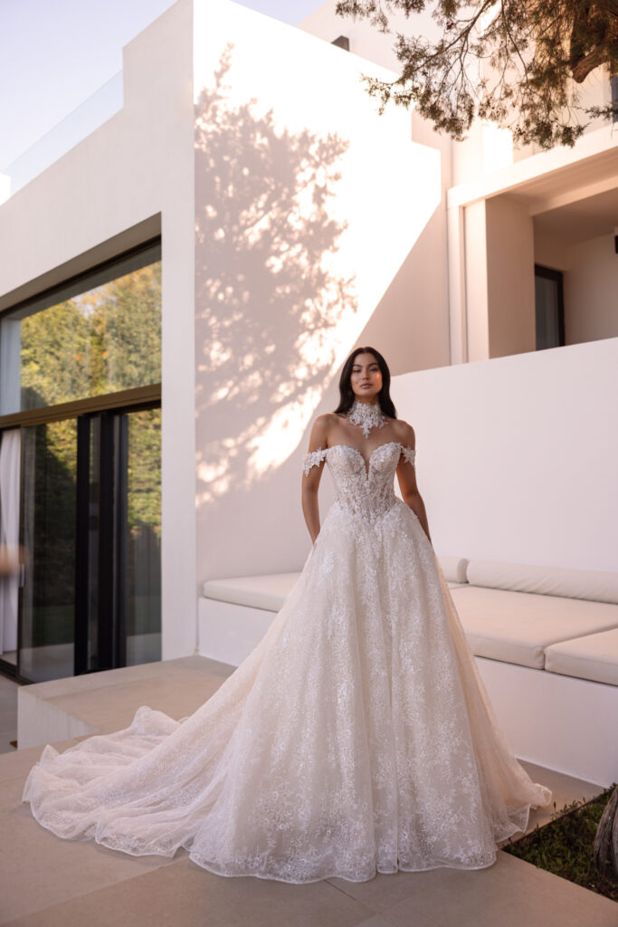 Prinzessinnen-Brautkleid aus Glittertüll mit tiefem Herzausschnitt, besticktem Korsett, abnehmbaren Off-Shoulder-Spitzenärmeln und Spitzen-Choker, Frontalansicht vor modernem Villenambiente.