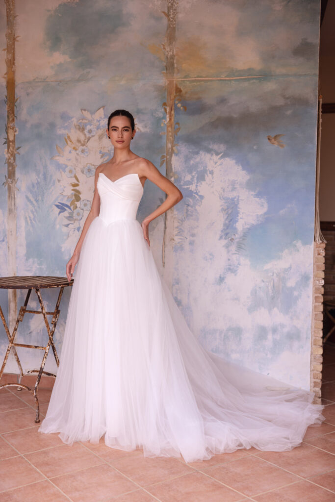 Braut in trägerlosem Brautkleid mit drapiertem Wickel-Oberteil und weit schwingendem Tüllrock, vor gemalter Wandkulisse.