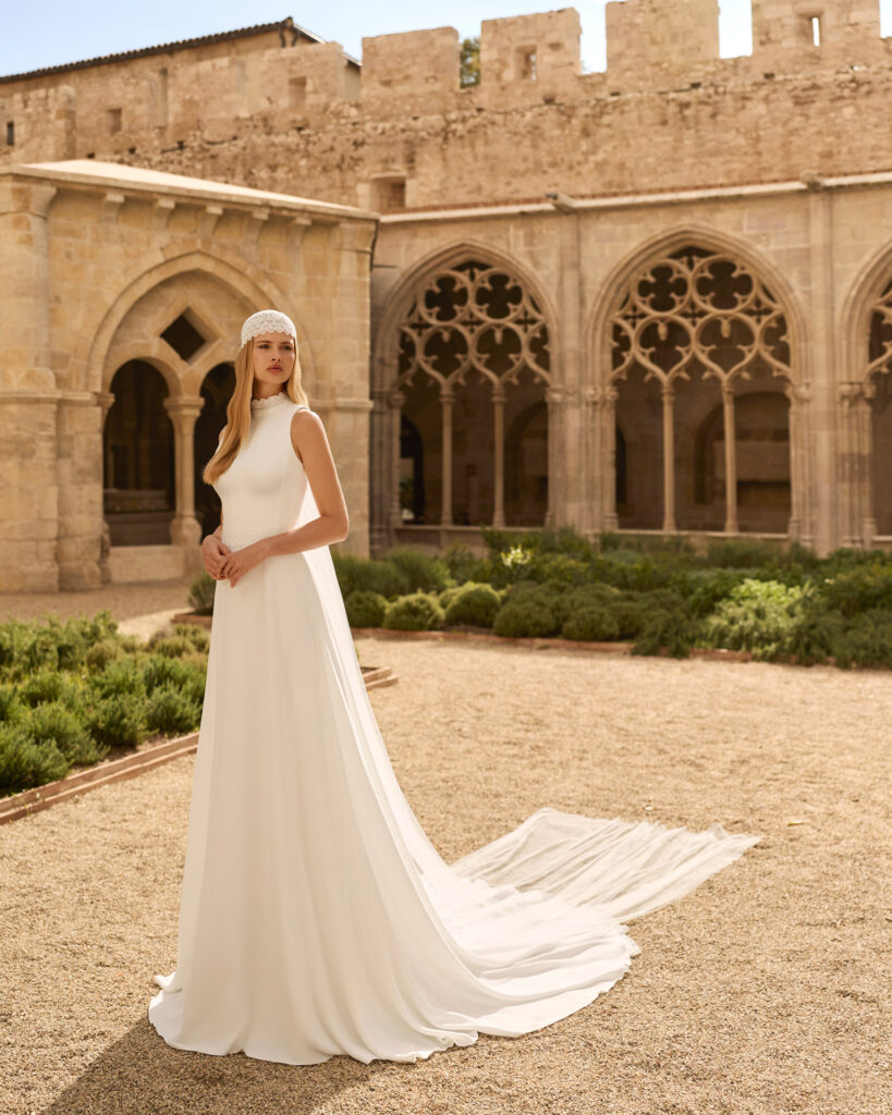 Braut in ärmellosem A-Linien-Brautkleid mit Stehkragen und Spitzenkäppchen vor historischer Burgmauer.